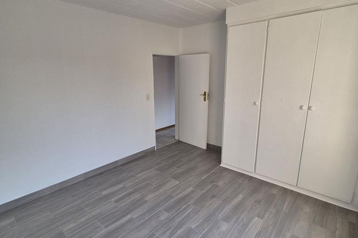 A bright, empty bedroom featuring modern grey wood-look tiled flooring and white built-in cupboards, offering a clean and neutral space.