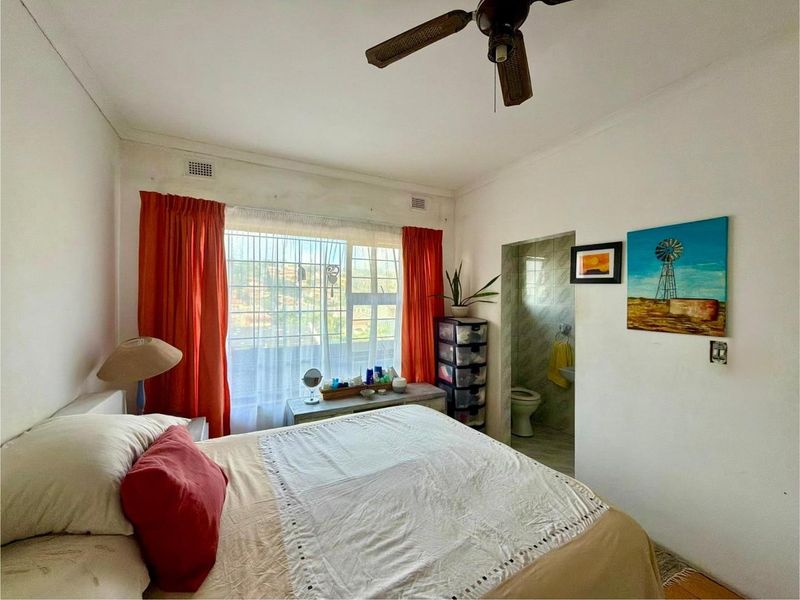 A bedroom featuring an en-suite bathroom, ceiling fan, and large window with natural light. The space requires cosmetic updates to improve its overall condition.