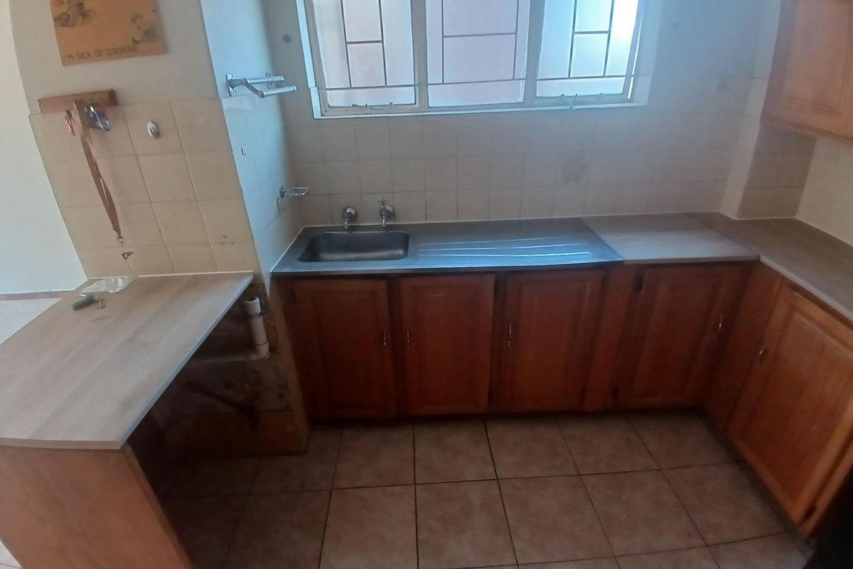 A dated kitchen featuring wooden cabinetry, laminate countertops, a stainless steel sink, and tiled flooring, requiring cosmetic updates and repairs to the cabinetry base.