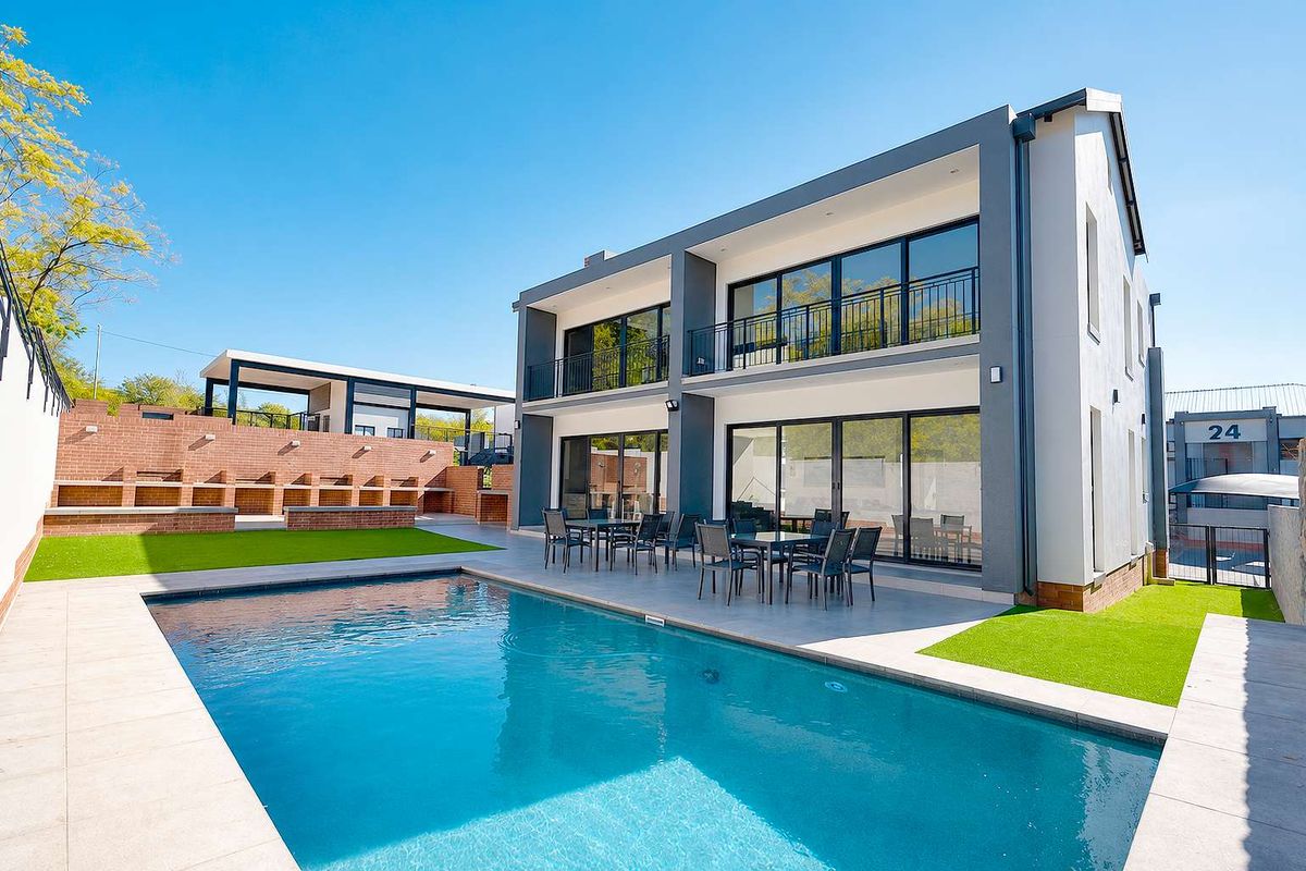 Modern two-story apartment exterior featuring a swimming pool, paved patio area, built-in brick braai, and upper-level balconies with glass sliding doors.