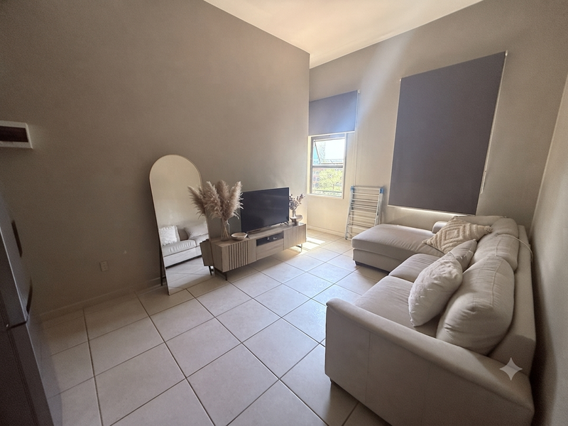 A bright living room featuring neutral ceramic floor tiles, large windows with roller blinds, and high ceilings, creating a spacious and airy atmosphere.