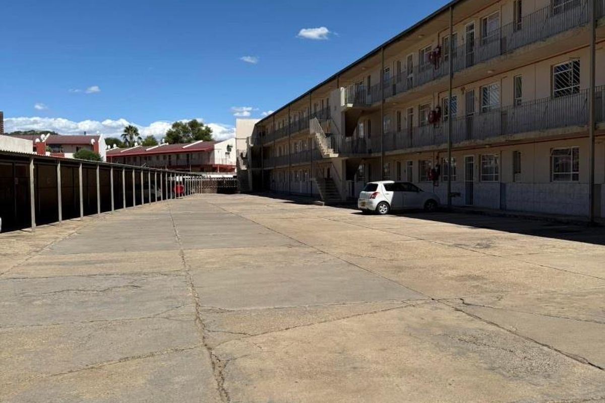 Exterior view of an apartment complex featuring a large concrete parking area and multi-level walkways with metal railings.