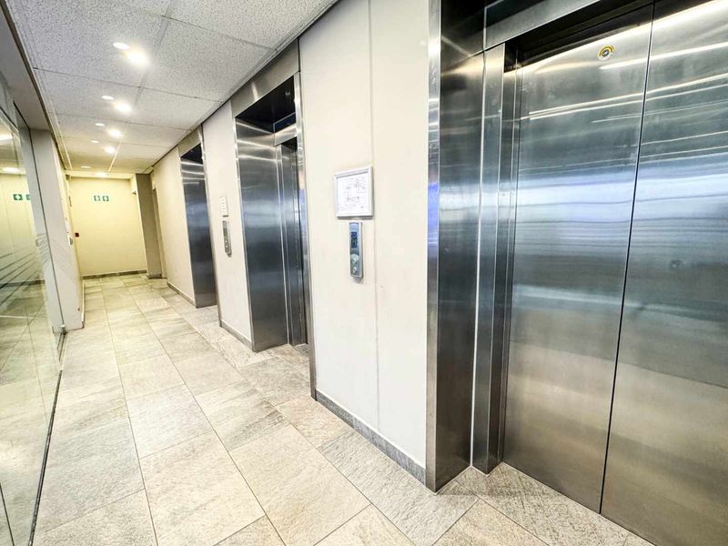 A clean and professional office building hallway featuring stainless steel elevator doors and neutral tiled flooring.