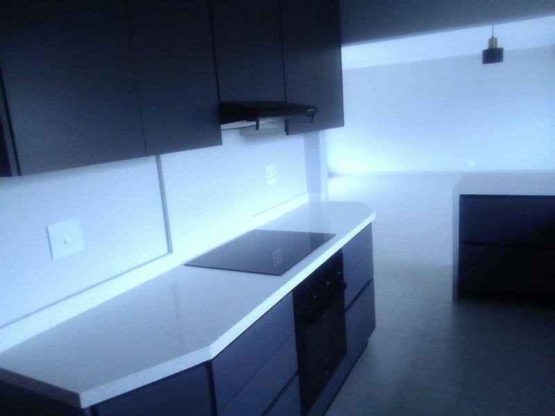 Modern kitchen featuring dark cabinetry, stone countertops, integrated electric hob, oven, and a matching kitchen island in an open-plan layout.