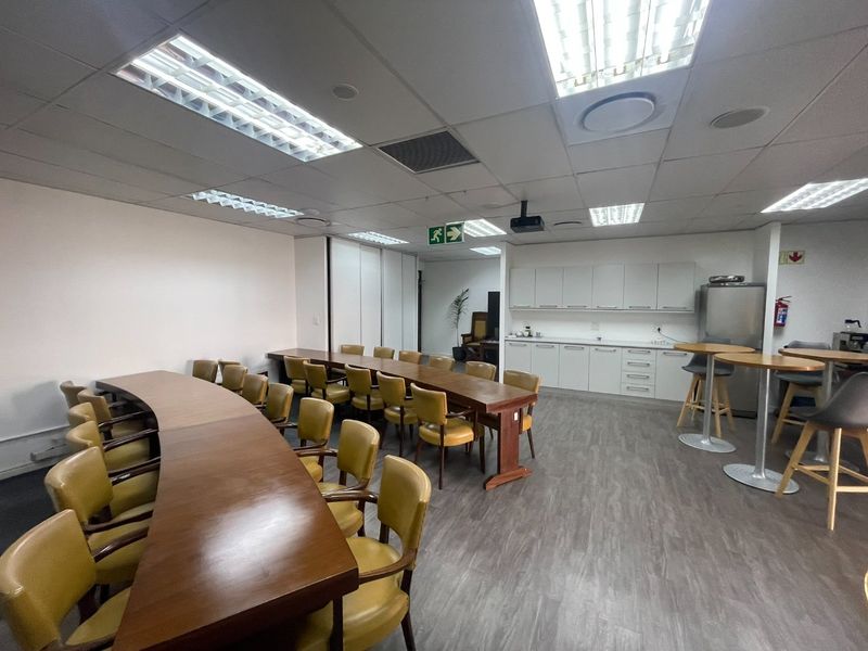 A functional office meeting room featuring a large curved conference table, built-in kitchenette cabinetry, and professional suspended ceiling lighting.