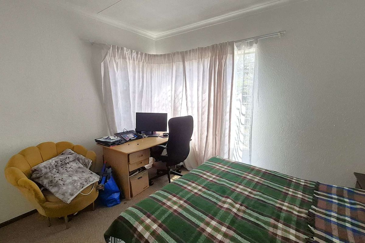 A bedroom featuring neutral walls, carpeted flooring, and large windows with curtains, providing a functional space for rest or work.