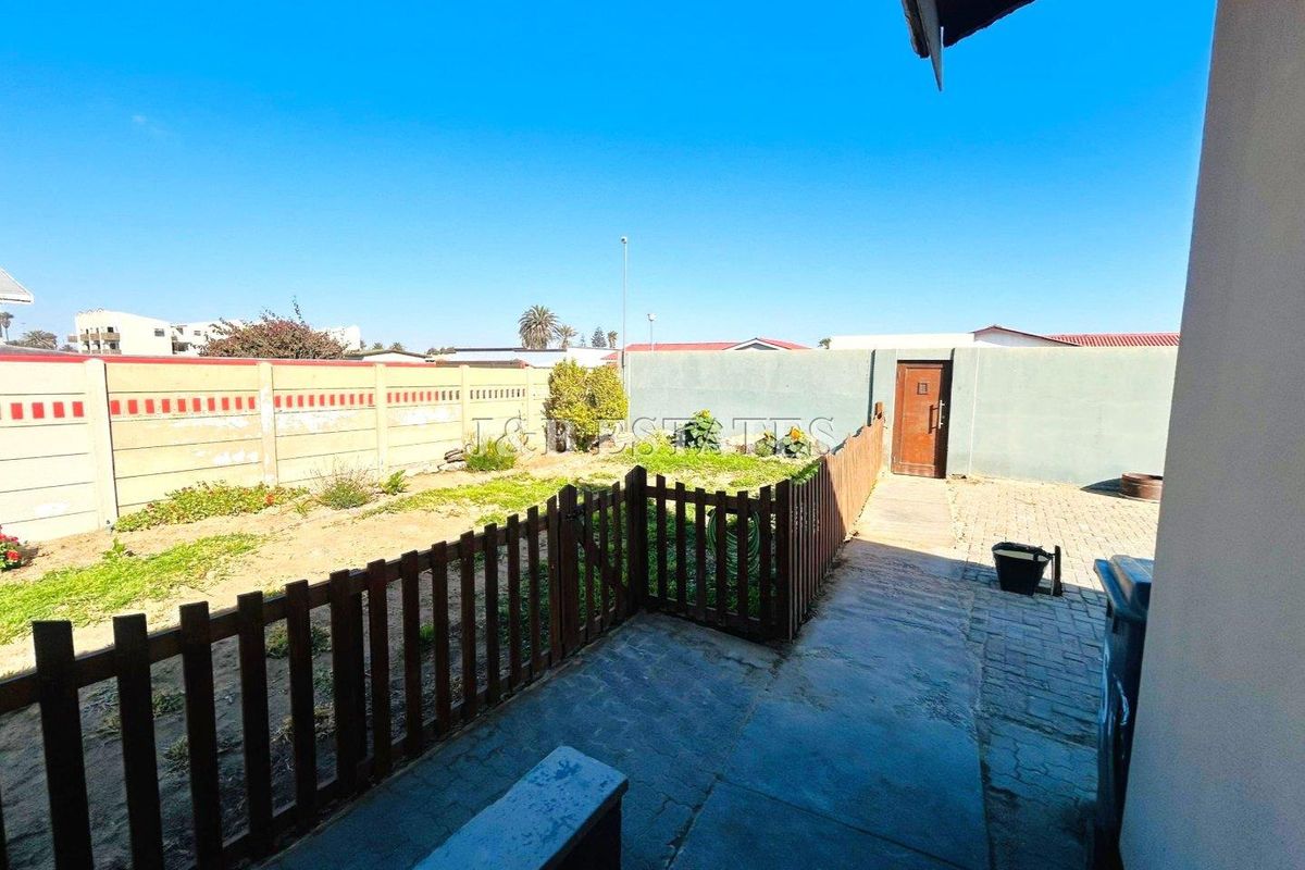A rear garden area featuring a paved walkway, a wooden picket fence, and a perimeter boundary wall with a rear access gate.