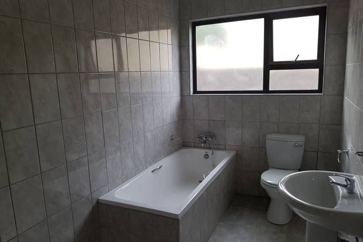 A functional bathroom featuring a built-in bathtub, pedestal basin, and toilet, finished with neutral wall-to-floor tiling and a window for natural light.