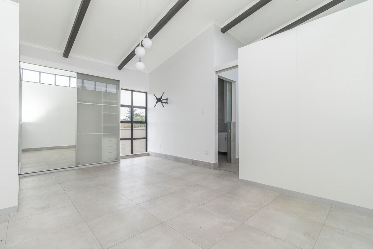 A bright, modern bedroom featuring vaulted ceilings with exposed beams, large tiled flooring, built-in sliding door wardrobes, and an en-suite bathroom entrance.