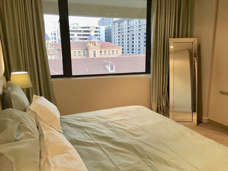 A well-lit bedroom featuring a large window with urban views, neutral wall finishes, and carpeted flooring.
