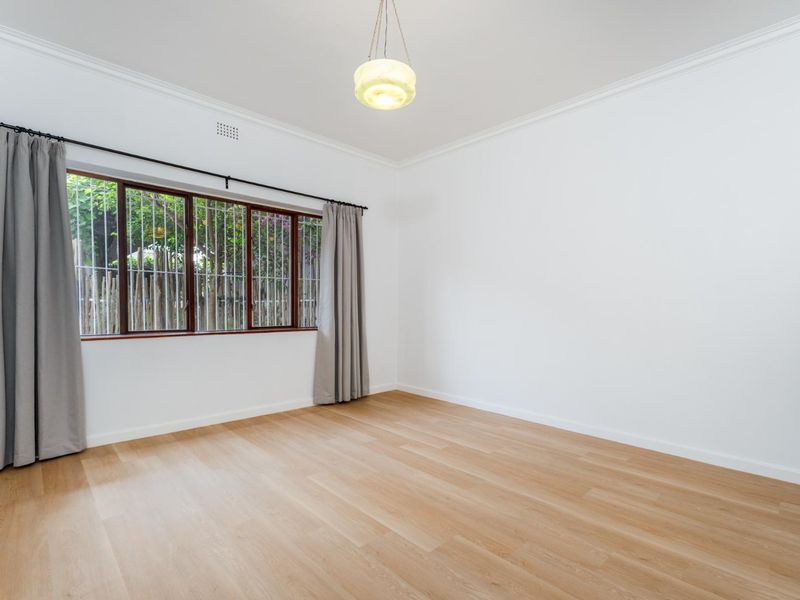 A bright, empty bedroom featuring light timber flooring, white walls, decorative cornice, and a window fitted with security bars.