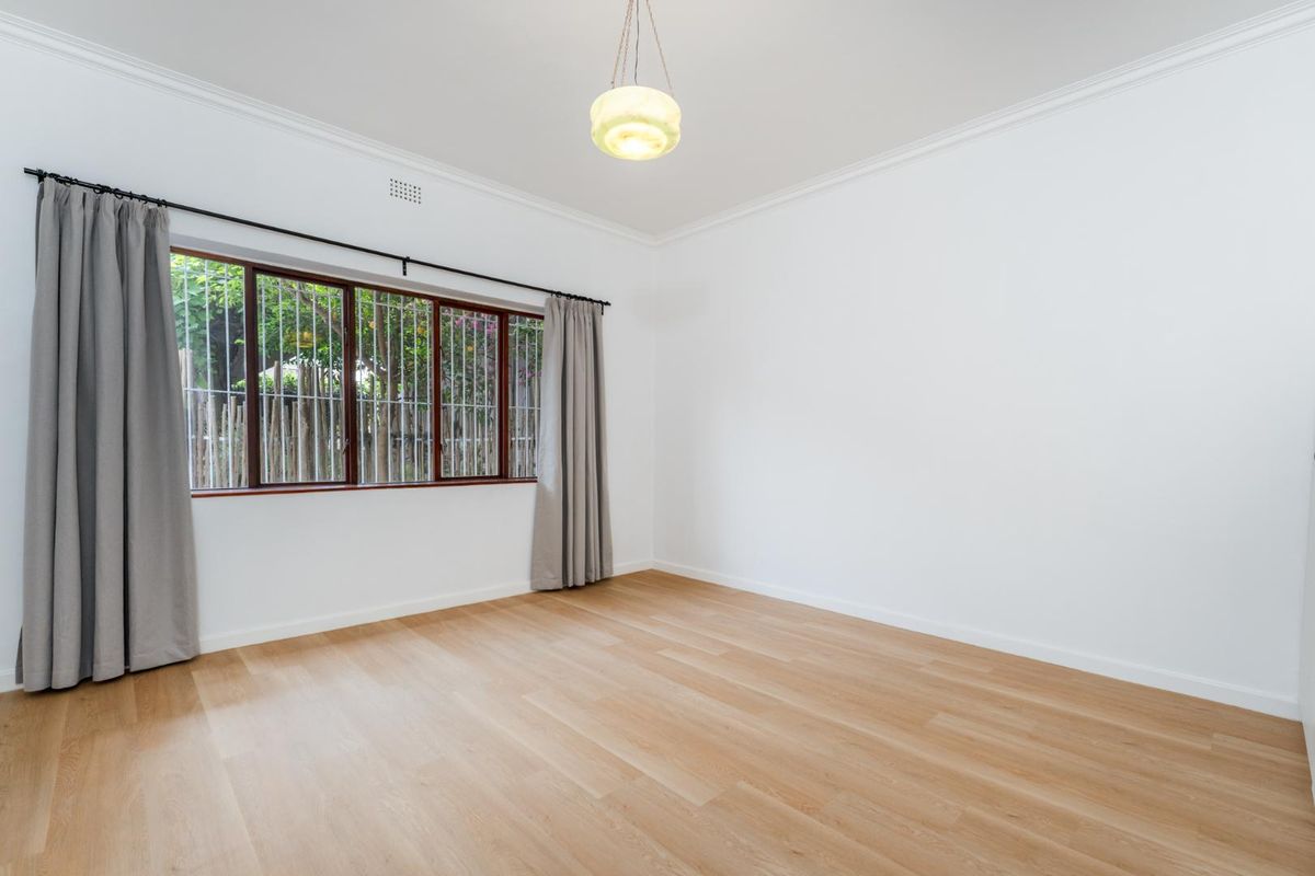 A bright, empty bedroom featuring light timber flooring, white walls, decorative cornice, and a window fitted with security bars.