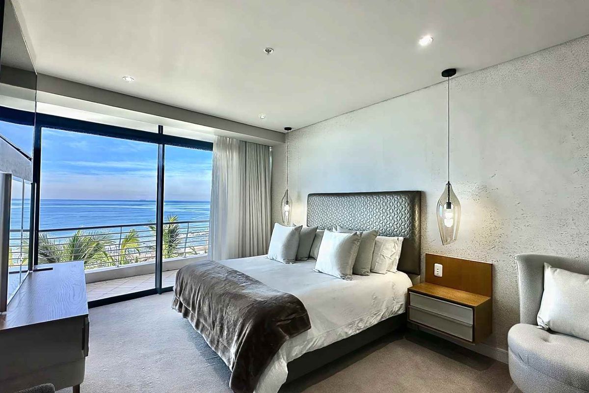 Modern bedroom featuring a textured feature wall, recessed ceiling lighting, and large sliding glass doors opening to a balcony with expansive ocean views.