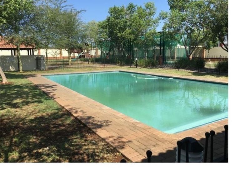 A communal swimming pool area featuring brick paving, surrounding metal fencing, and established garden landscaping within the apartment complex.