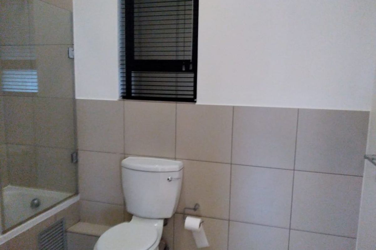 A functional bathroom featuring a toilet, glass shower enclosure, and neutral wall tiling with a window for natural light.