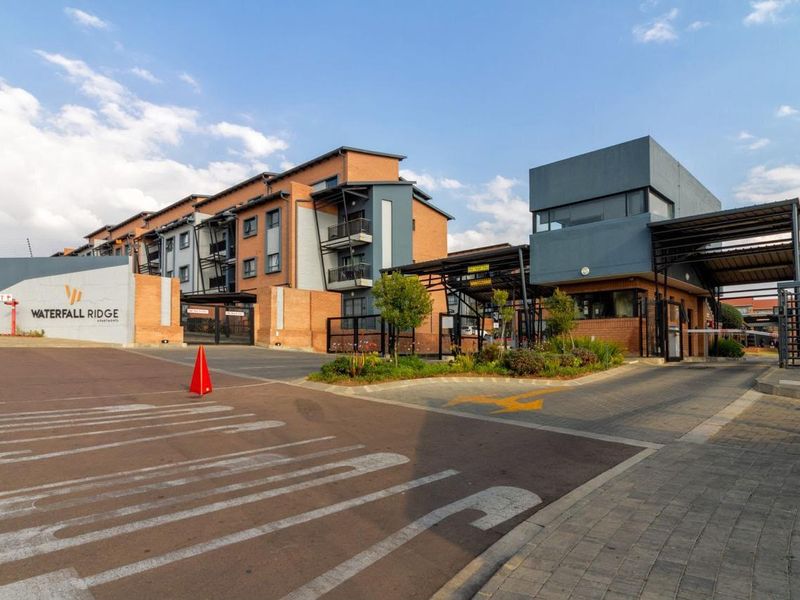 The main entrance and security gatehouse of Waterfall Ridge Apartments, featuring modern brick architecture, paved access, and secure perimeter fencing.