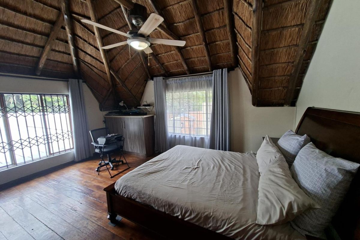 Spacious bedroom featuring a high vaulted thatch ceiling, exposed timber beams, wooden flooring, and large windows with security bars.