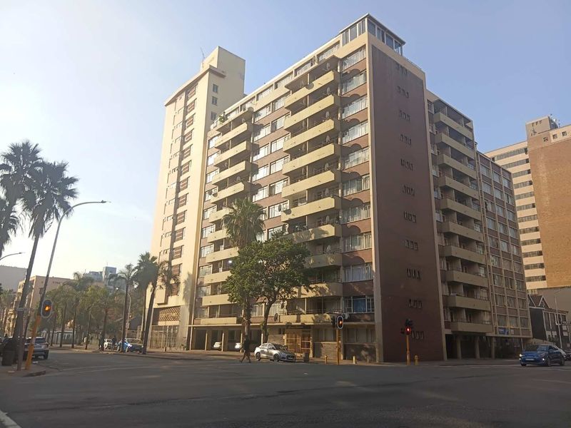 Street view of a multi-story apartment building featuring multiple balconies and a ground-level parking area.