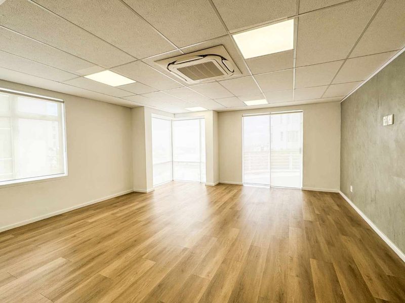 A bright, modern, and empty office space featuring laminate flooring, a suspended ceiling with recessed lighting, and large windows providing ample natural light.