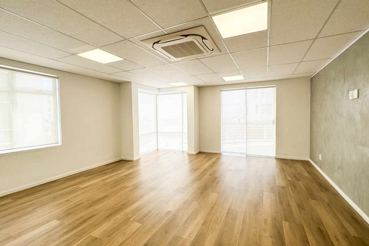 A bright, modern, and empty office space featuring laminate flooring, a suspended ceiling with recessed lighting, and large windows providing ample natural light.