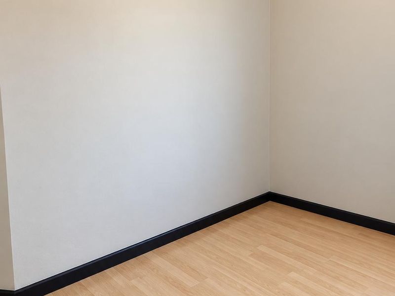 A clean, empty bedroom featuring light-toned laminate flooring, contrasting dark skirting boards, and neutral wall finishes.