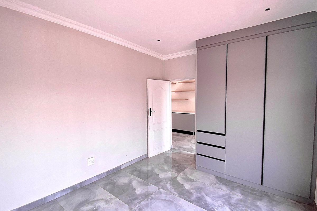 A bright bedroom featuring modern grey floor tiles, built-in cupboards, and crown molding, with an open doorway leading to an adjacent room.
