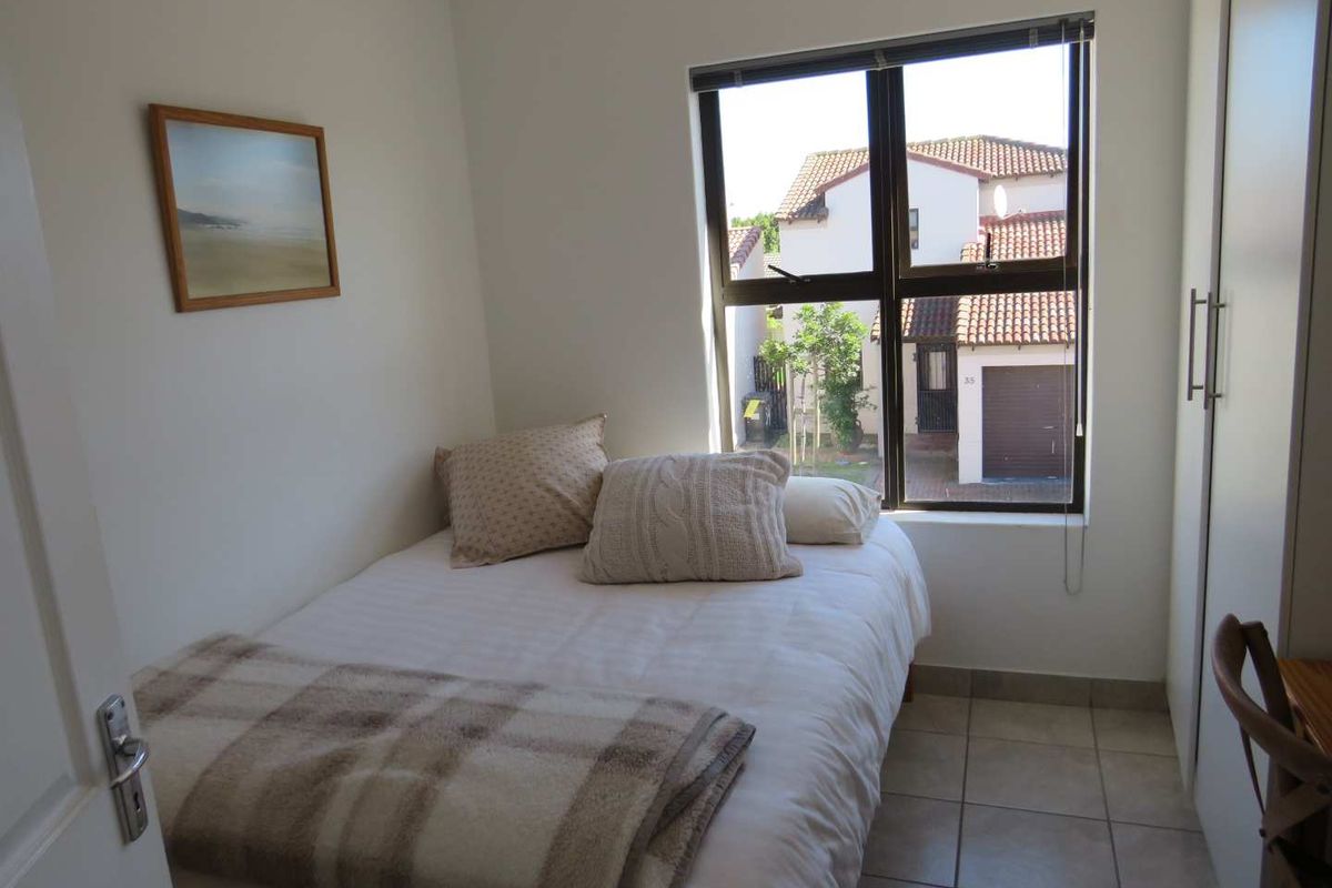 A compact bedroom featuring tiled flooring, a built-in wardrobe, and a window providing natural light and a view of the surrounding residential area.