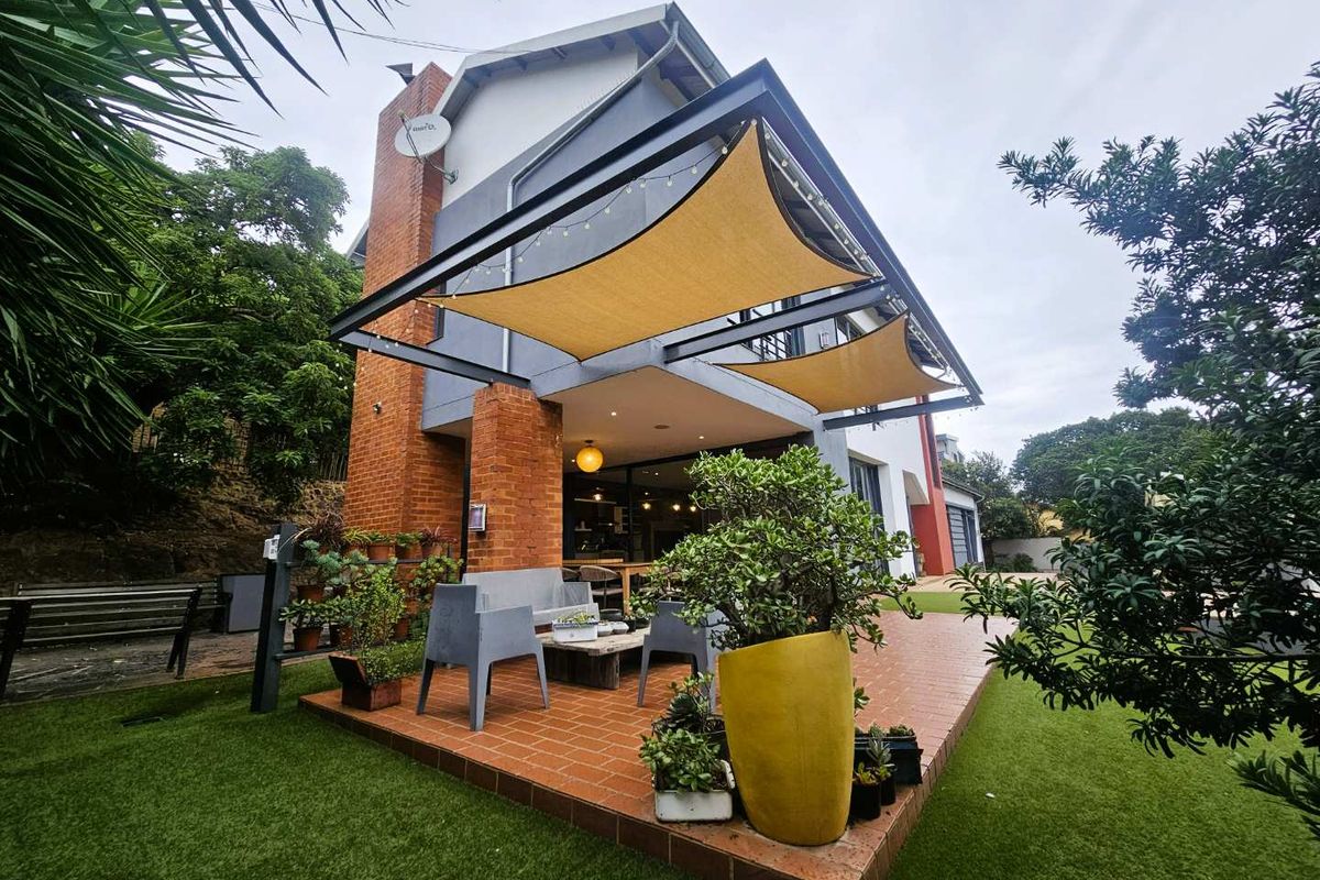 A covered rear patio featuring a brick base, steel structural frame, and shade sails, overlooking a landscaped garden area.