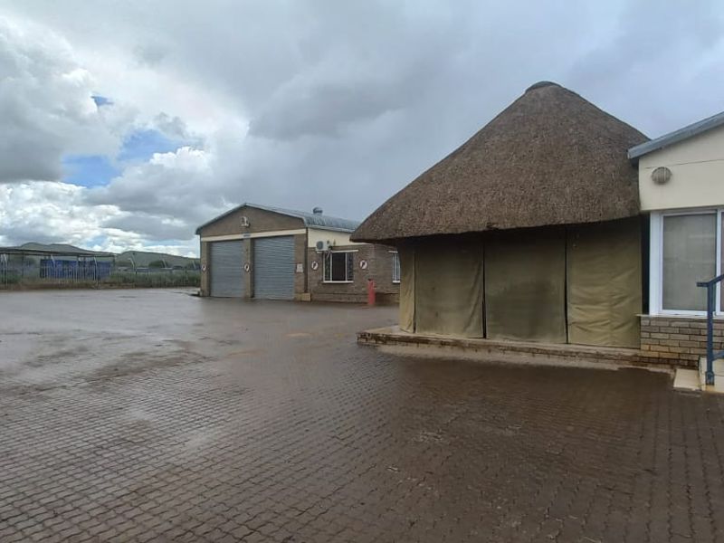 Industrial property exterior featuring a large paved yard, roller shutter garage doors, and a building with a traditional thatched roof section.