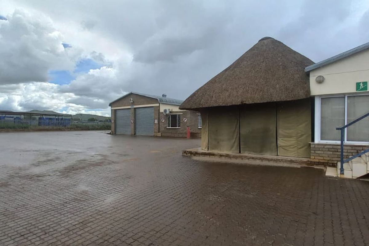 Industrial property exterior featuring a large paved yard, roller shutter garage doors, and a building with a traditional thatched roof section.