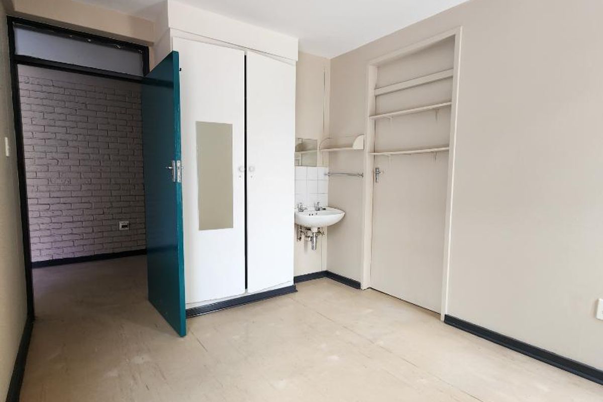 An empty bedroom featuring a built-in wardrobe, wall-mounted basin, and built-in shelving, with neutral walls and vinyl flooring.