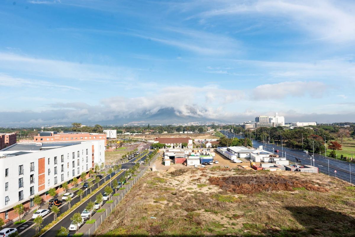 Elevated aerial view showcasing the surrounding neighbourhood, nearby apartment complex, and distant mountain landscape under a clear blue sky.