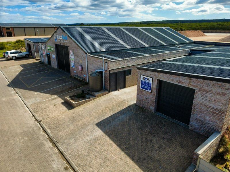 Aerial view of a commercial industrial property featuring brick construction, corrugated metal roofing, roller shutter doors, and a paved parking area.
