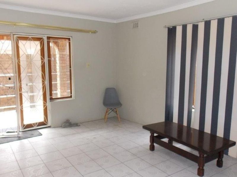 A spacious, neutrally painted living area featuring tiled flooring, a sliding door with a security gate, and vertical blinds.
