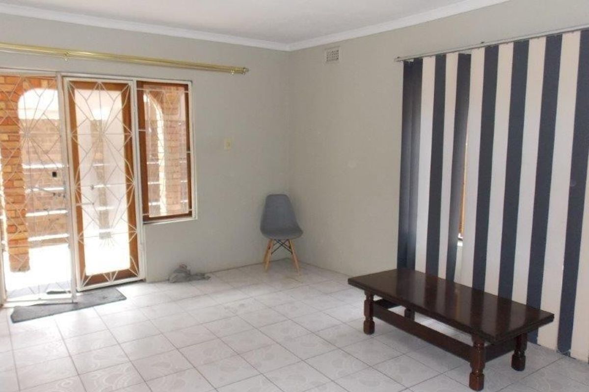 A spacious, neutrally painted living area featuring tiled flooring, a sliding door with a security gate, and vertical blinds.