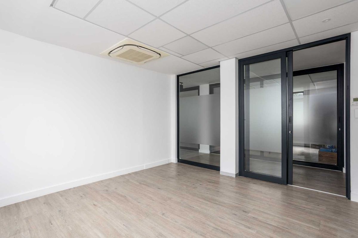 A modern, empty office space featuring laminate flooring, a suspended ceiling with integrated air conditioning, and sleek sliding glass partitions.