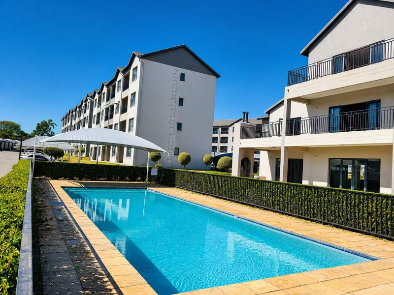 A well-maintained communal swimming pool area featuring a shade sail, paved decking, and neat perimeter hedging within the apartment complex.