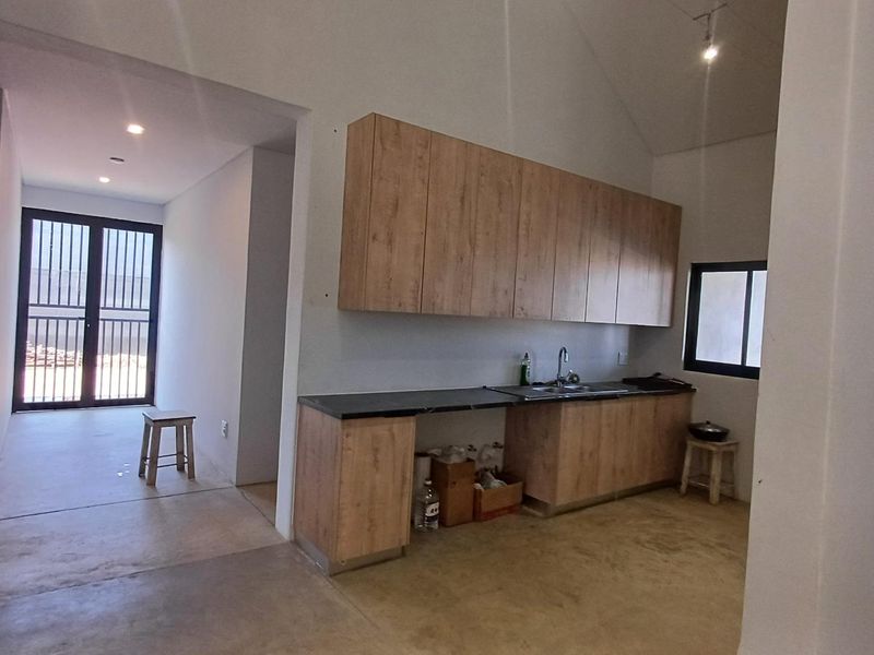 A functional kitchen area featuring wood-look cabinetry, dark countertops, and a stainless steel sink, set within an open-plan space with vaulted ceilings.
