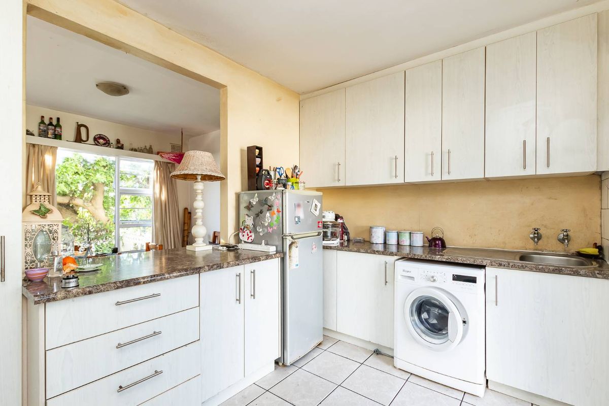 A functional kitchen featuring white cabinetry, laminate countertops, a stainless steel sink, and space for a washing machine, opening into an adjacent living area.