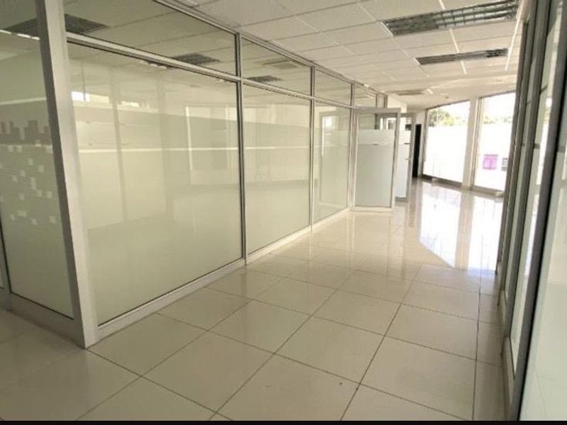 A bright, vacant office space featuring modern glass partitions, tiled flooring, and a suspended ceiling with recessed lighting.