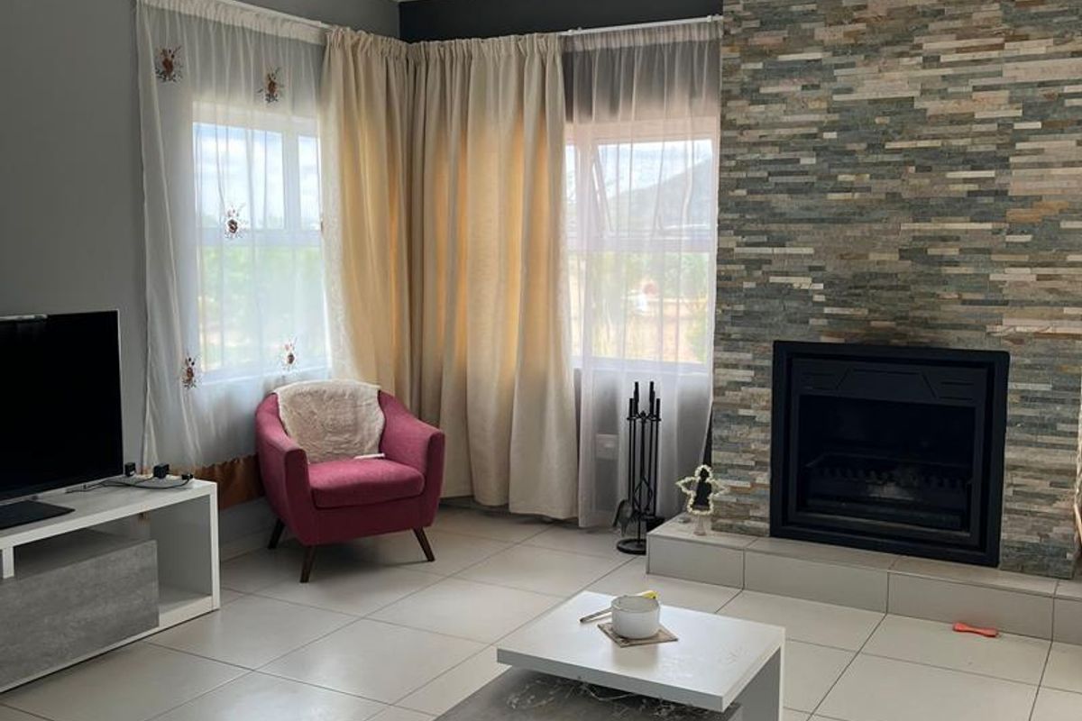 A comfortable living room featuring a prominent stone-clad fireplace, neutral tiled flooring, and large windows allowing for natural light.