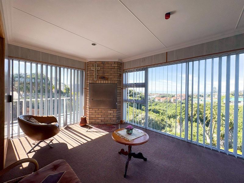 A sunlit living room featuring a built-in brick fireplace, expansive windows with vertical blinds, and scenic elevated views.