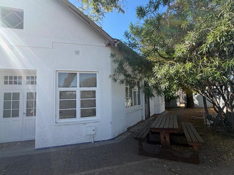 Rear exterior view of the property featuring a white rendered facade, paved patio area, and mature trees providing shade.