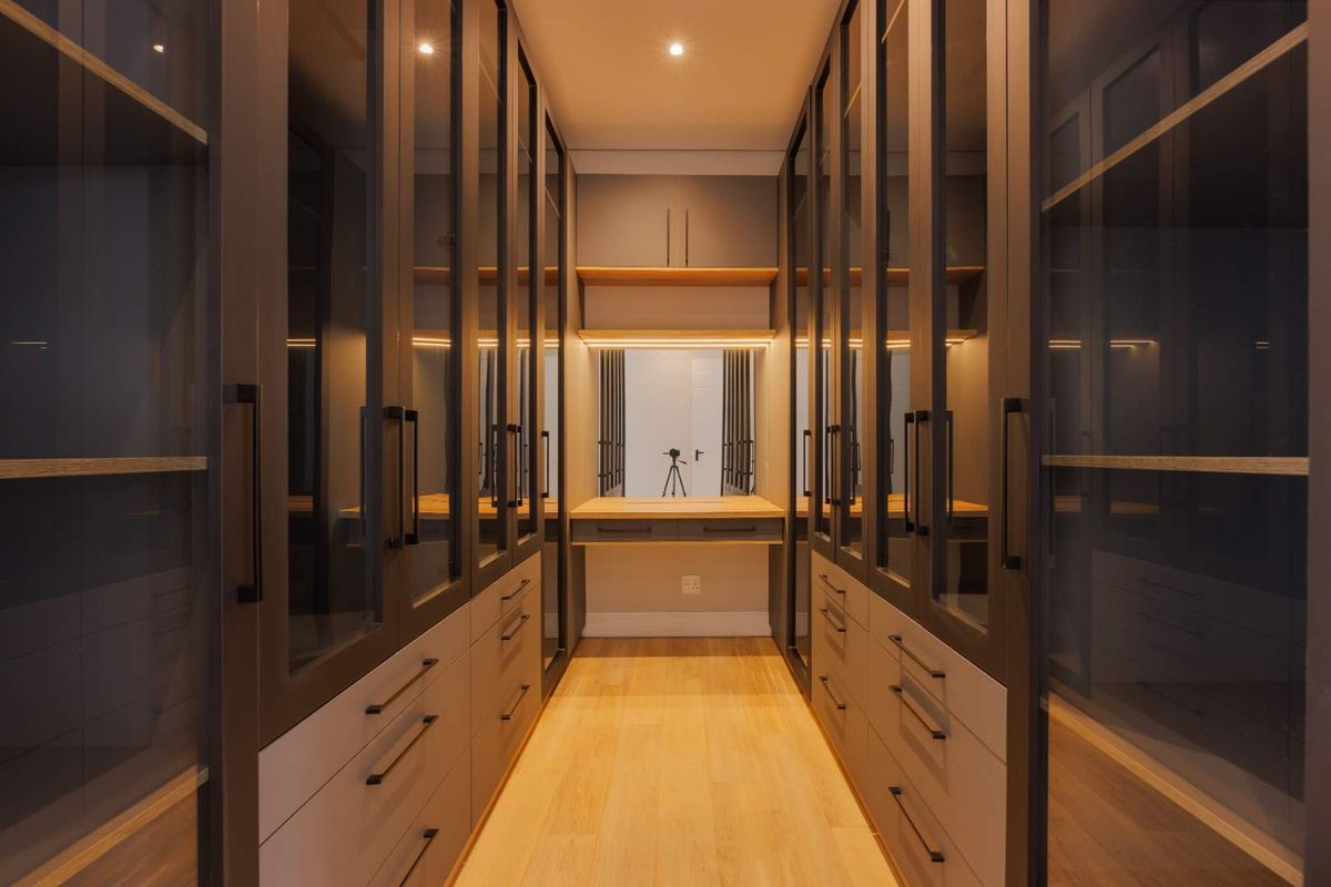 A modern walk-in dressing room featuring extensive floor-to-ceiling glass-fronted cabinetry, integrated lighting, and a central vanity station with light wood flooring.