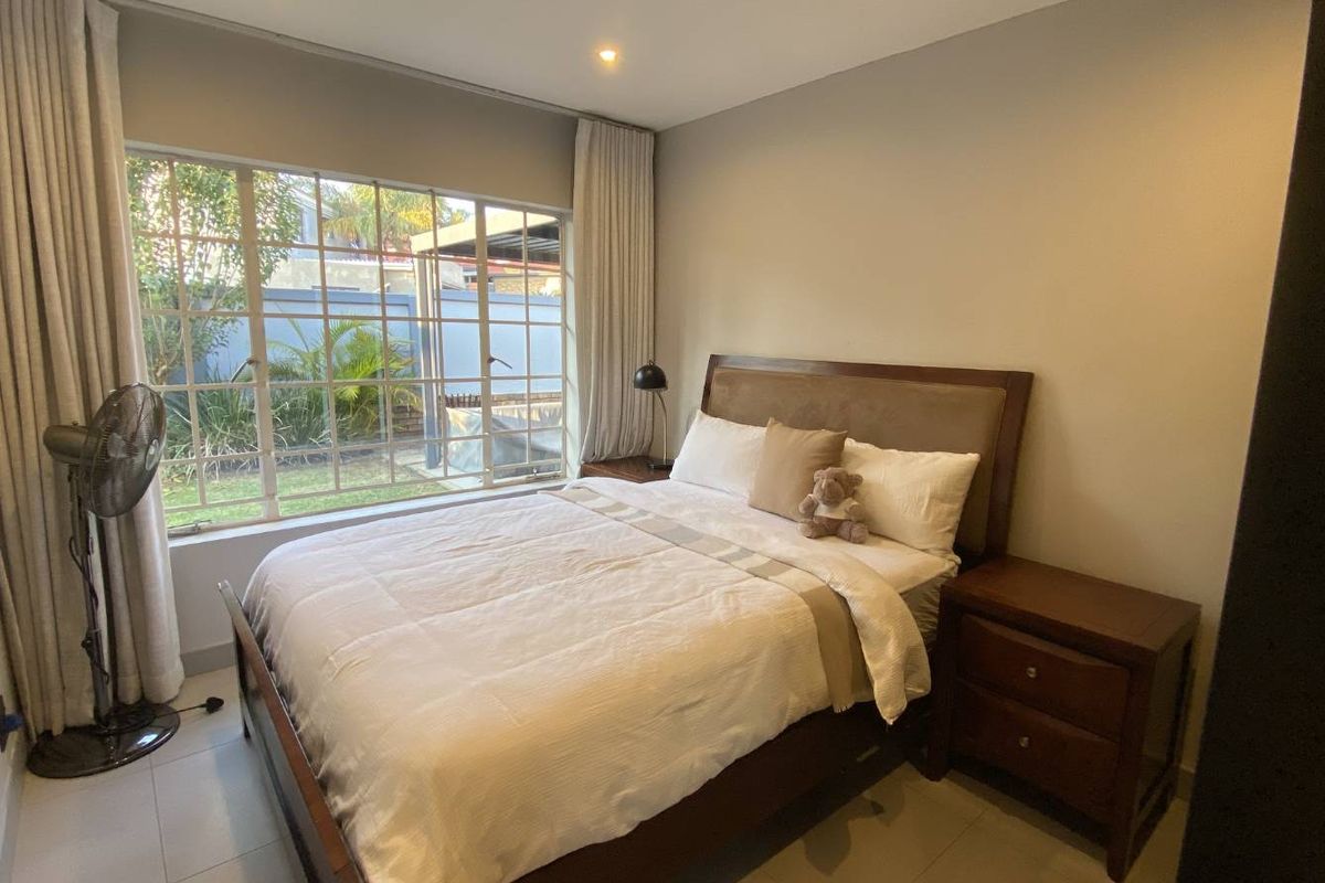 A well-lit bedroom featuring neutral tiled flooring, recessed ceiling lighting, and a large window overlooking the garden area.