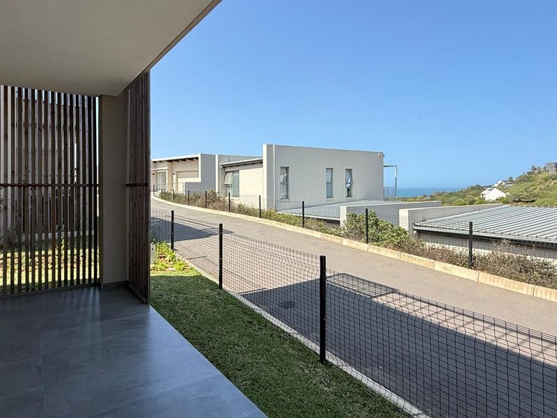 A modern covered patio featuring sleek tiled flooring, timber slat privacy screening, and recessed lighting, overlooking a paved driveway and coastal landscape.