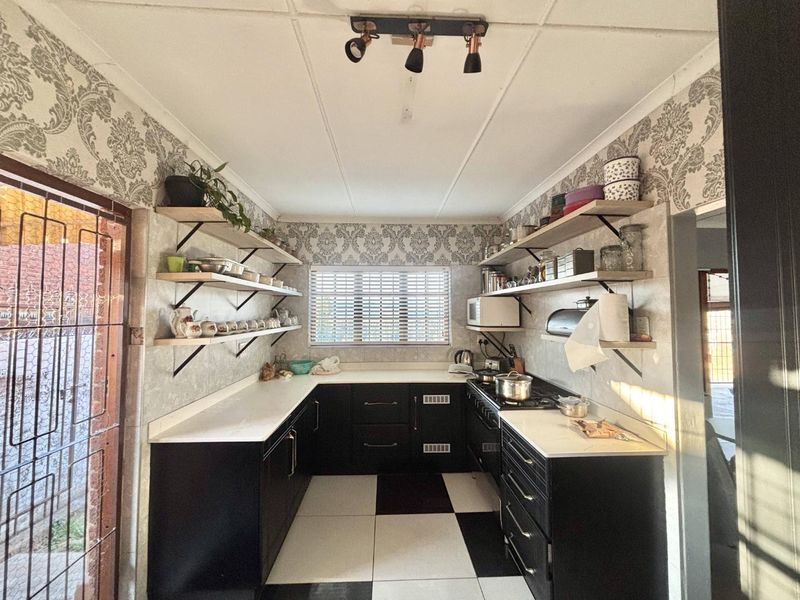 A functional kitchen featuring black cabinetry, white countertops, open shelving, and a gas hob, finished with patterned wallpaper and checkered floor tiles.