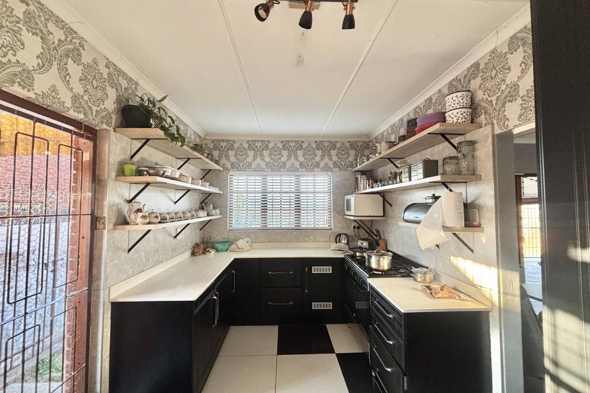 A functional kitchen featuring black cabinetry, white countertops, open shelving, and a gas hob, finished with patterned wallpaper and checkered floor tiles.