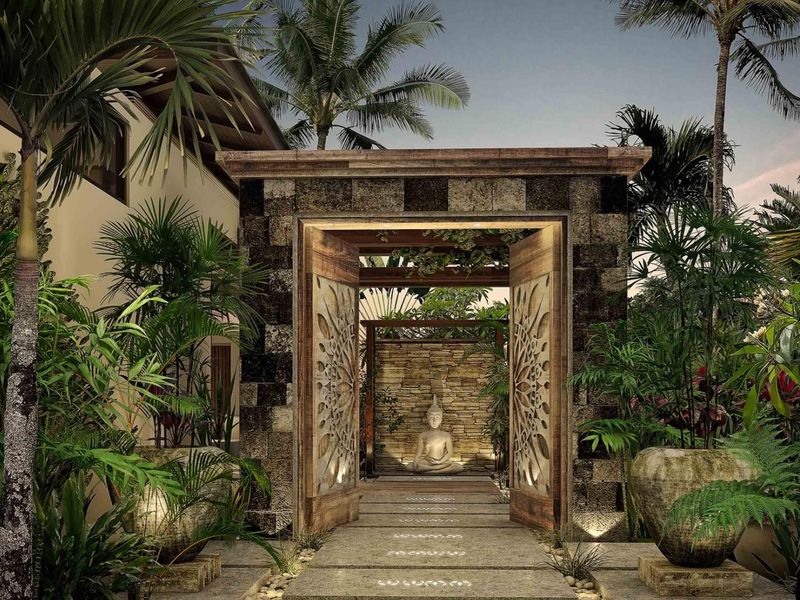 A beautifully landscaped front entrance featuring a stone masonry gate, intricate carved wooden doors, and a stone pathway leading to a serene garden area.