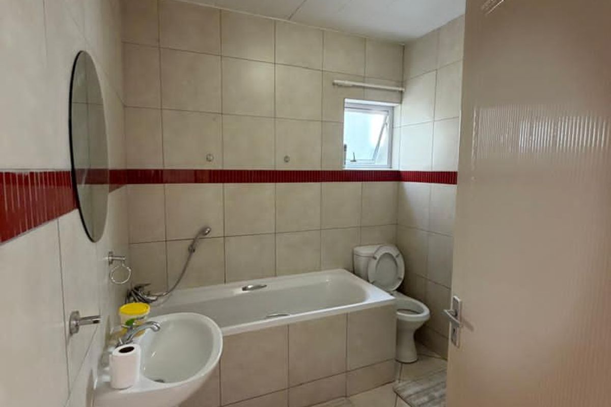 A functional bathroom featuring a pedestal sink, a bathtub with a shower mixer, and a toilet, finished with neutral wall tiles and a red accent.